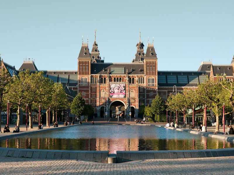 The Rijksmuseum in Amsterdam, featuring a tree-lined path and a reflecting pool in the foreground, under a clear blue sky.