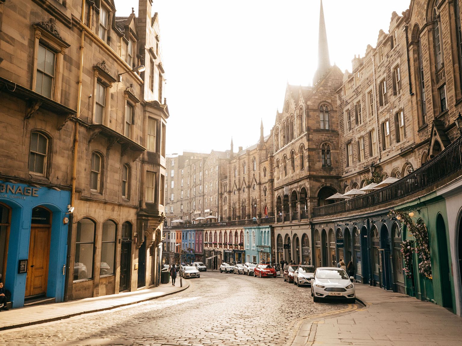 Street scene with historic buildings, colorful facades, and parked cars in sunlight, capturing an architectural blend of styles and curved road.