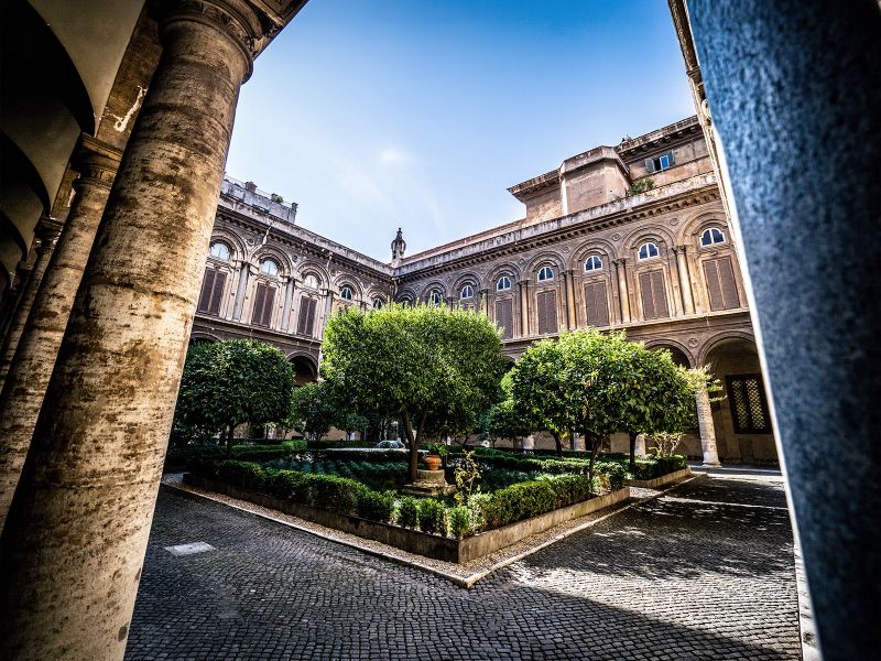 Historic courtyard with arched windows, symmetrical garden, and a fountain, framed by stone columns under a clear blue sky.