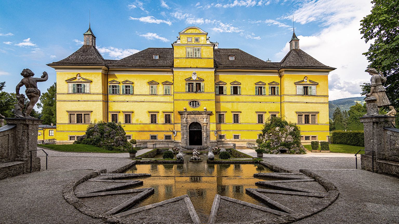 Bright yellow palace with ornate architecture, surrounded by statues and a star-shaped fountain, under a blue sky with clouds.