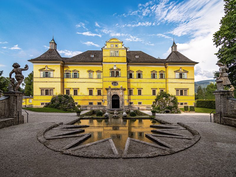 Bright yellow palace with ornate architecture, surrounded by statues and a star-shaped fountain, under a blue sky with clouds.