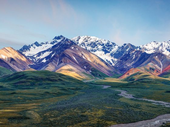 Majestic mountain landscape with snow-capped peaks, colorful slopes, and a lush green valley under a clear blue sky.