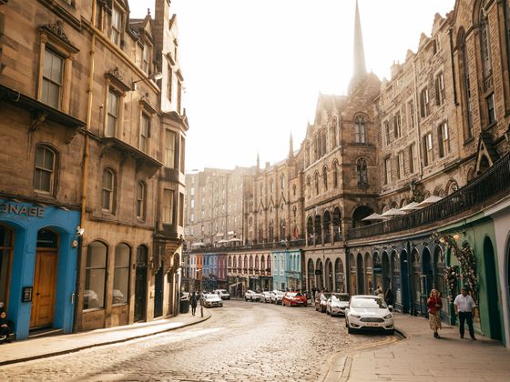 Sunlit cobblestone street lined with historic buildings and colorful shopfronts, cars parked along the road, and people walking.
