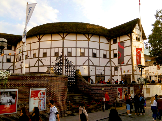 The Globe Theatre, a circular thatched building with timber framing, surrounded by visitors and flags, on a sunny day.