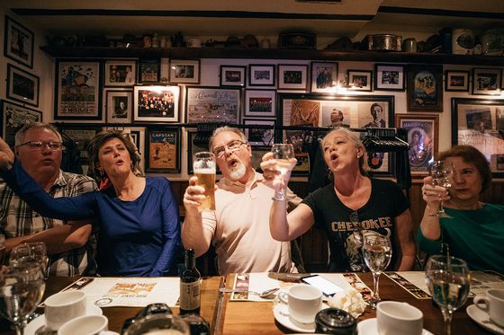 Five people seated at a restaurant table, energetically singing and raising glasses, with walls adorned with numerous framed pictures.