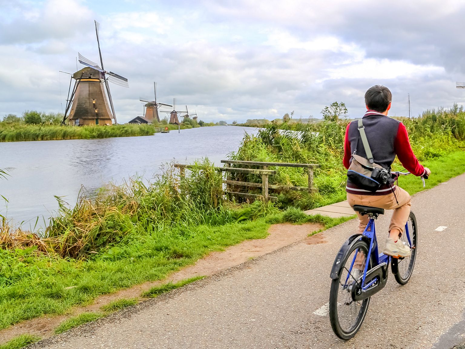 Person cycling on a path beside a river with windmills in the background under a cloudy sky.