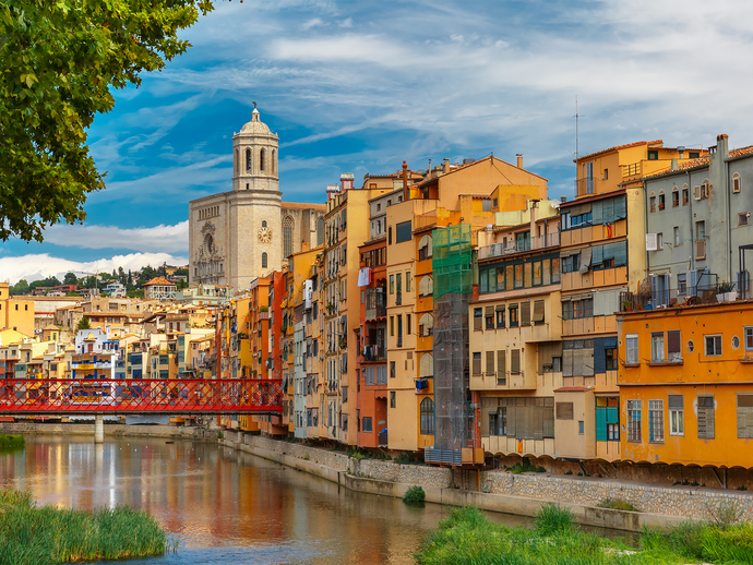 Colorful riverside buildings in Girona, Spain, with a view of the Cathedral of Saint Mary. A red bridge crosses the Onyar River below a blue sky.