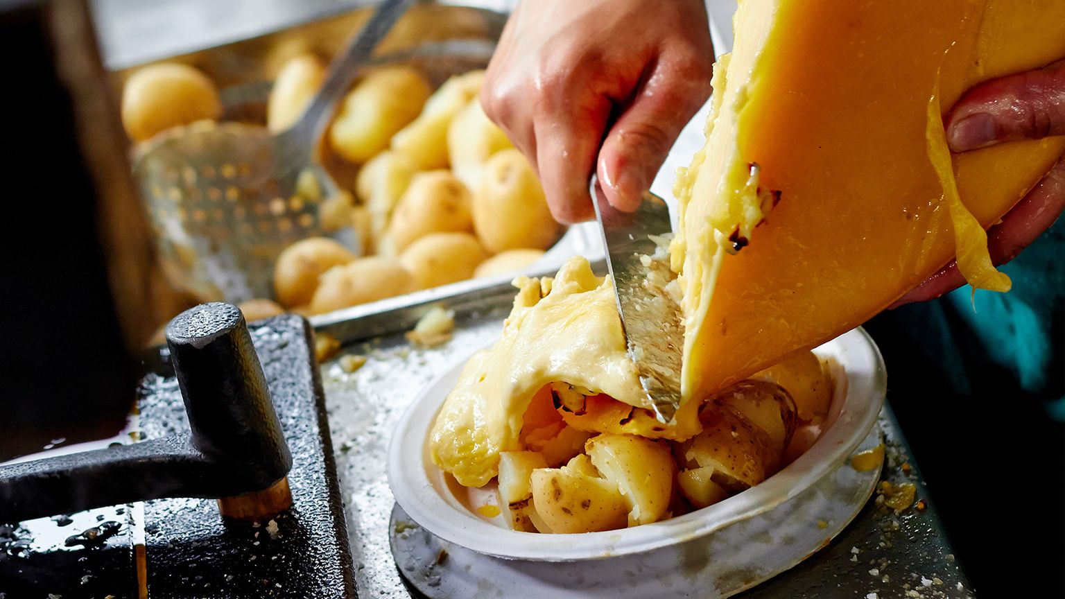 Hands slicing melted cheese onto boiled potatoes, using a large knife. A skimmer with additional potatoes is visible in the background.