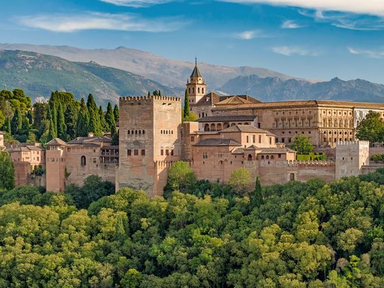 The Alhambra, a historic fortified palace with towers, surrounded by lush greenery and backed by distant mountains under a blue sky.