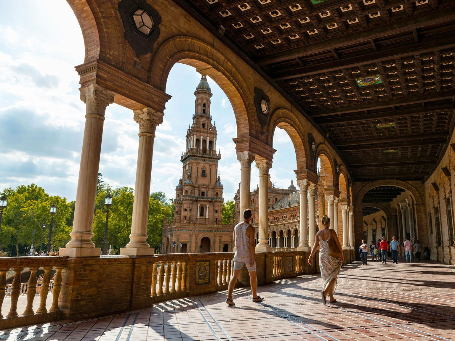 People walking under arched colonnade at Plaza de España, Seville, with a view of the central tower and vibrant sky in the background.