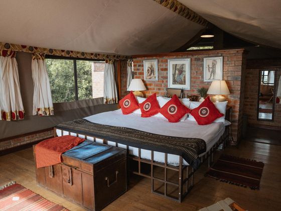 Cozy bedroom with a large bed, red accent pillows, brick wall, framed art, and large windows with nature views. Warm lighting and wooden decor.
