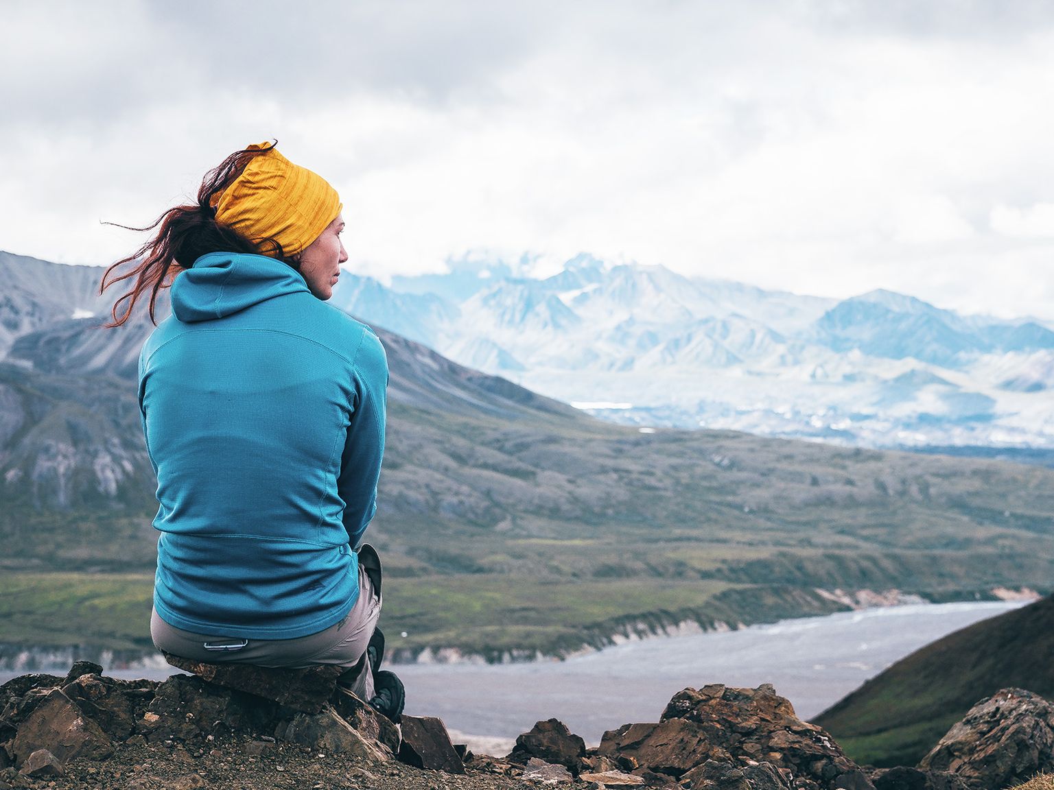 Person in a blue jacket and yellow headscarf sitting on a rocky hill, overlooking a vast mountain landscape under a cloudy sky.