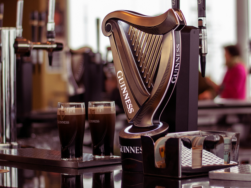 Guinness beer taps with a harp design on a bar counter alongside two poured glasses and a metal tray.