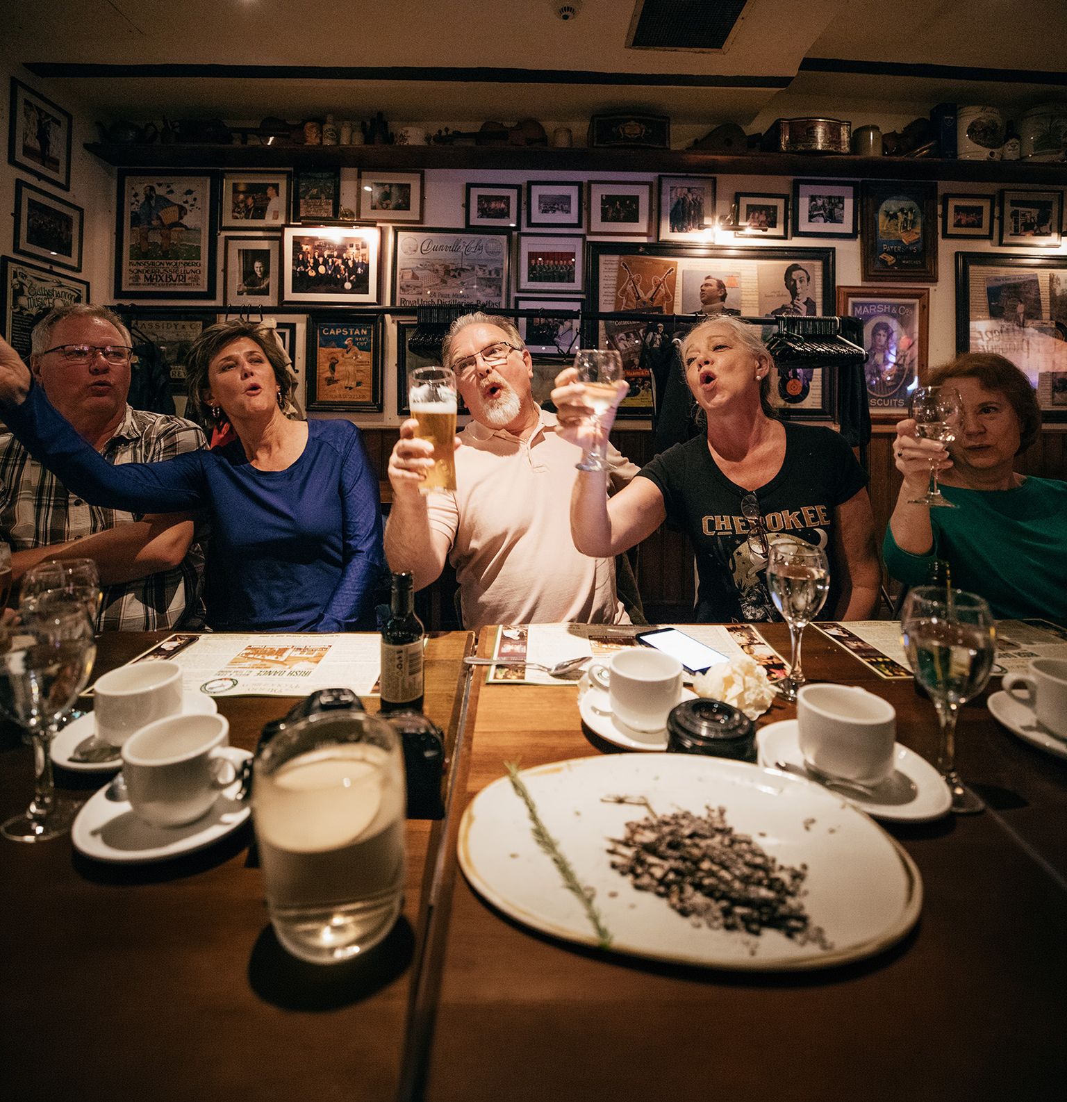 A group of people at a lively table in a restaurant, raising glasses and singing, surrounded by a warm, decoratively framed wall.