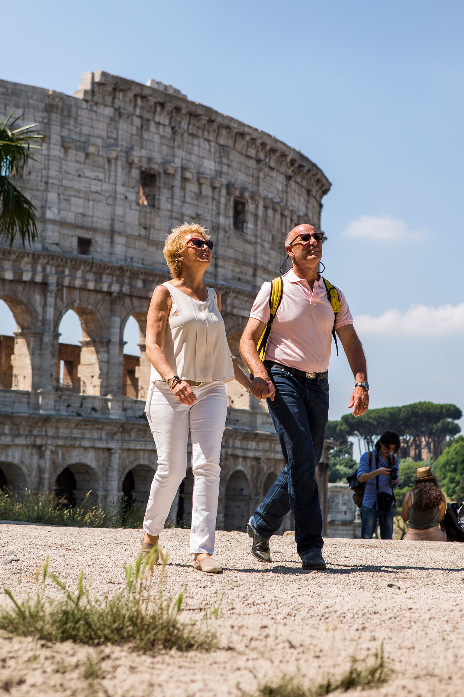 A smiling couple walks near the Colosseum in Rome on a sunny day, with a few people in the background.