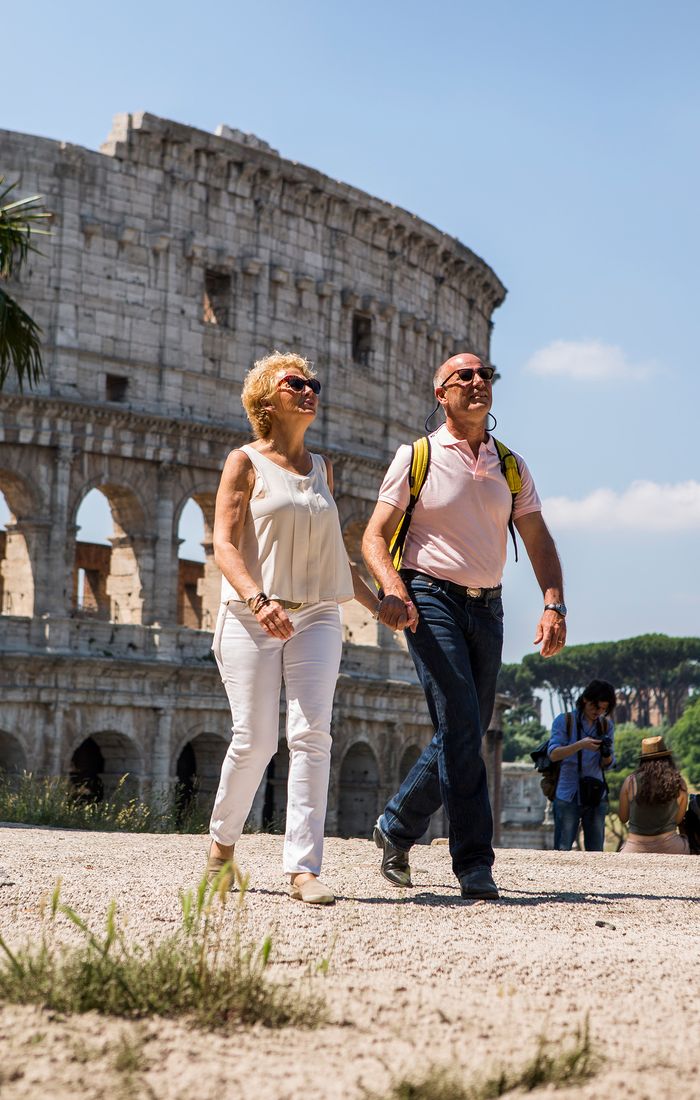 A smiling couple walks near the Colosseum in Rome on a sunny day, with a few people in the background.