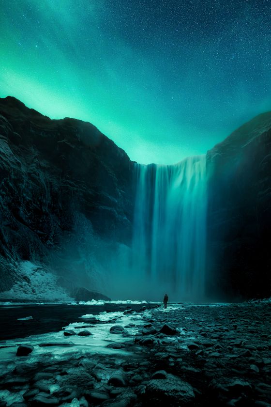 A person stands near a waterfall under a starry night sky with green auroras, surrounded by towering cliffs and rocky terrain.