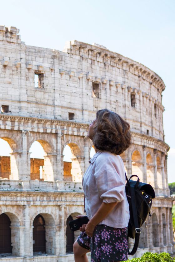 A person with a backpack stands looking at the Colosseum in Rome, with arches and ancient stone walls visible in the background.