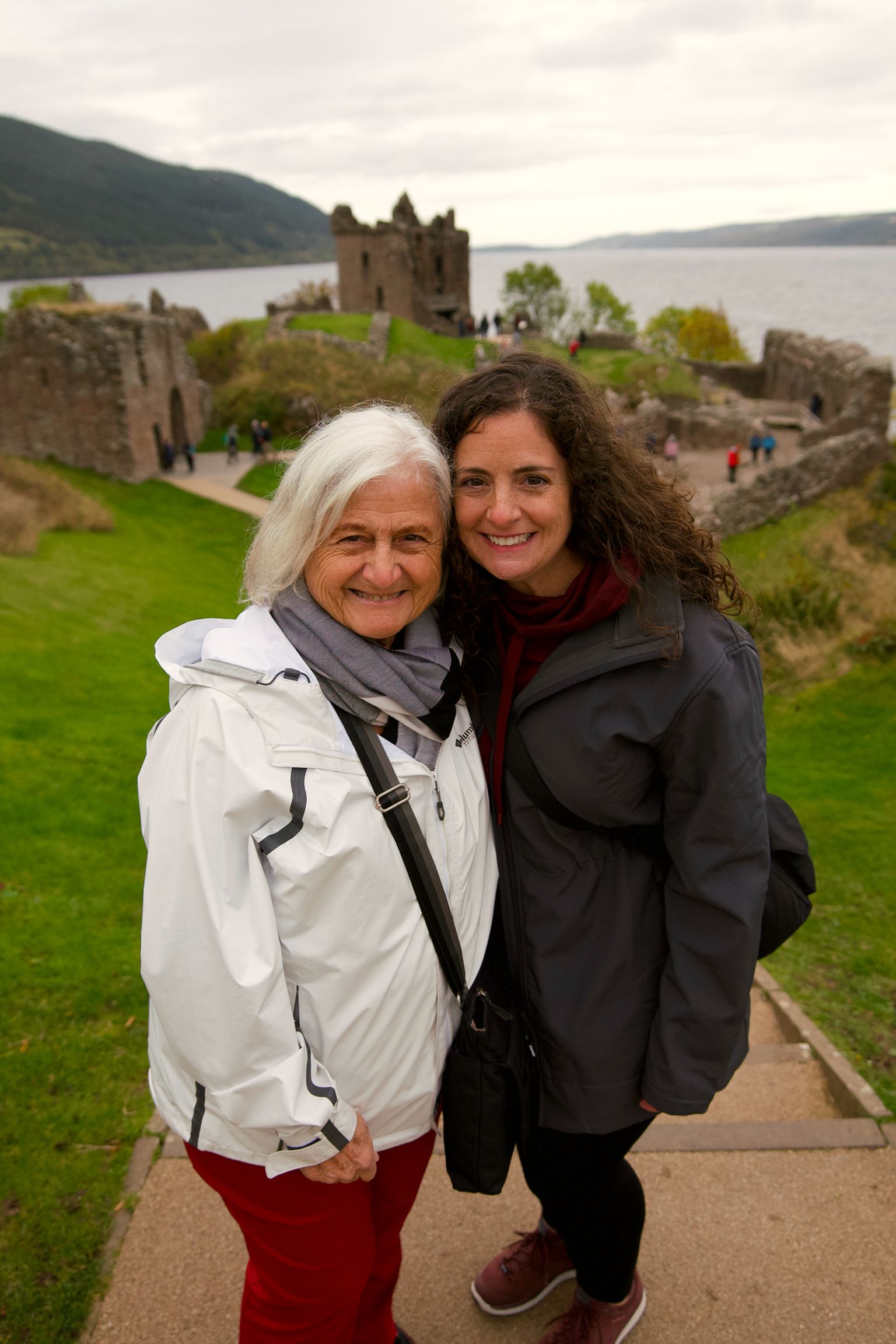 Two women smiling on a path overlooking a historic stone castle by a lake, with green hills in the background.