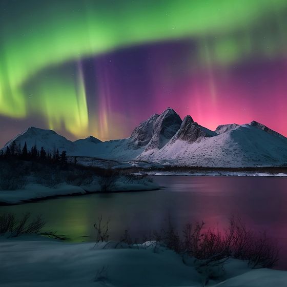Vibrant aurora borealis lights up a snowy mountain landscape in shades of green and pink, reflecting on a calm lake below.