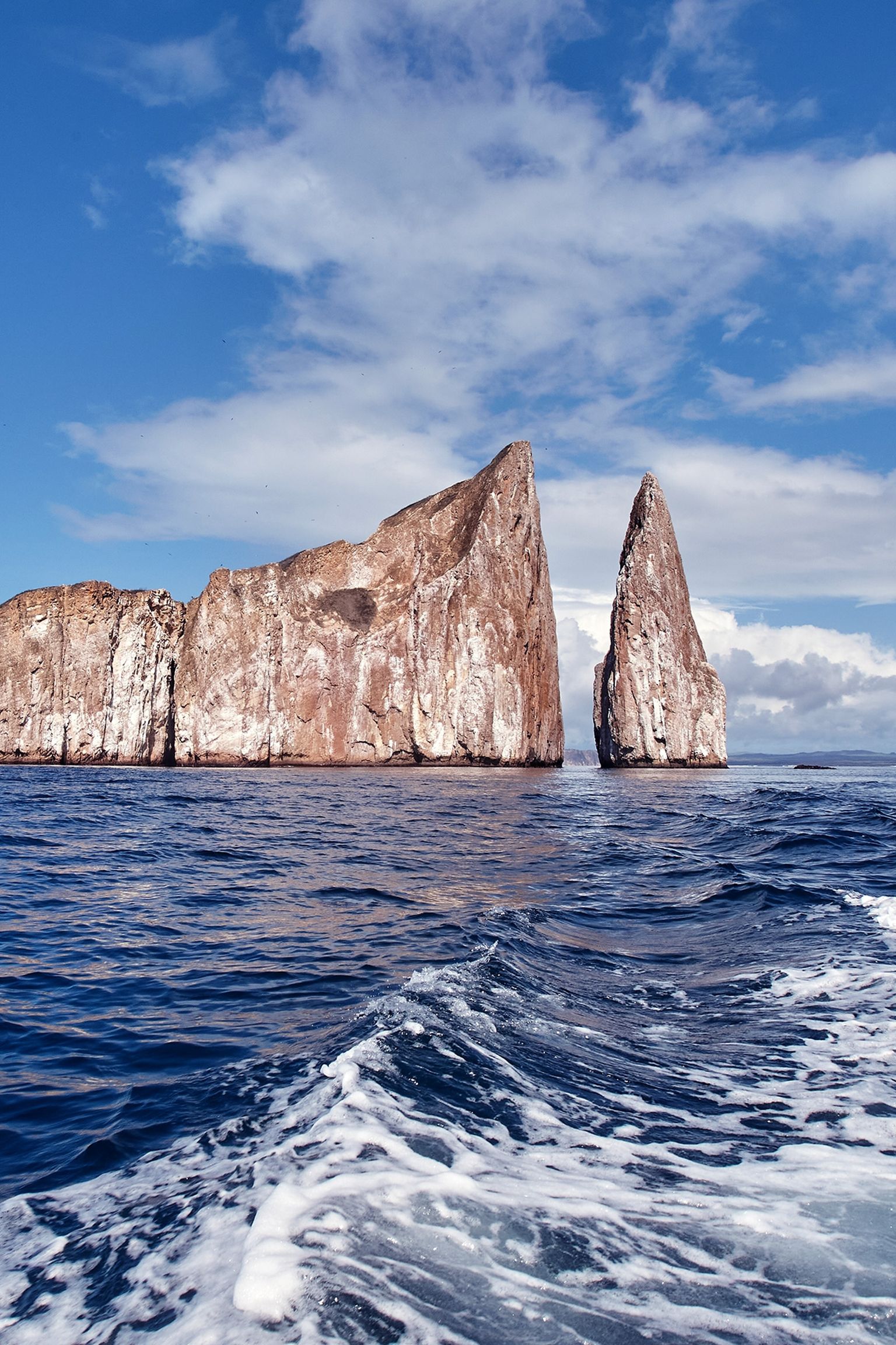 Dramatic rock formations rise from the ocean under a blue sky with clouds, waves in the foreground create a sense of movement.