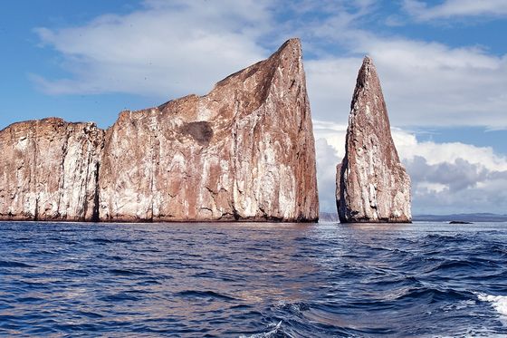 Dramatic rock formations rise from the ocean under a blue sky with clouds, waves in the foreground create a sense of movement.