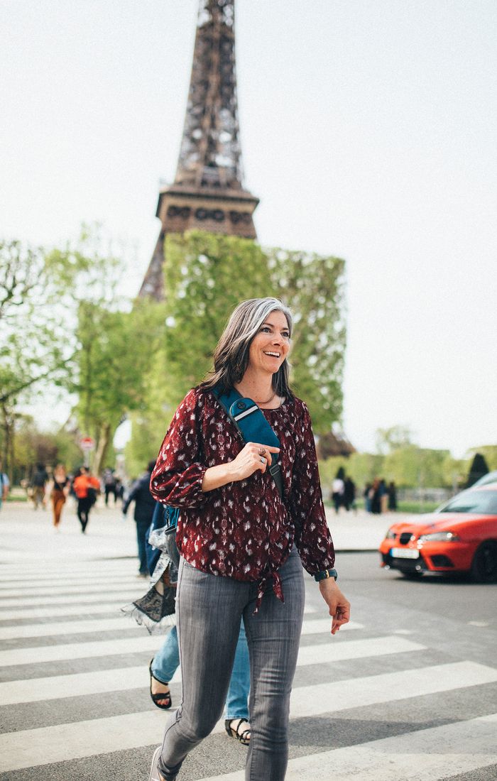 Smiling woman in a patterned blouse crossing a street with the Eiffel Tower in the background.