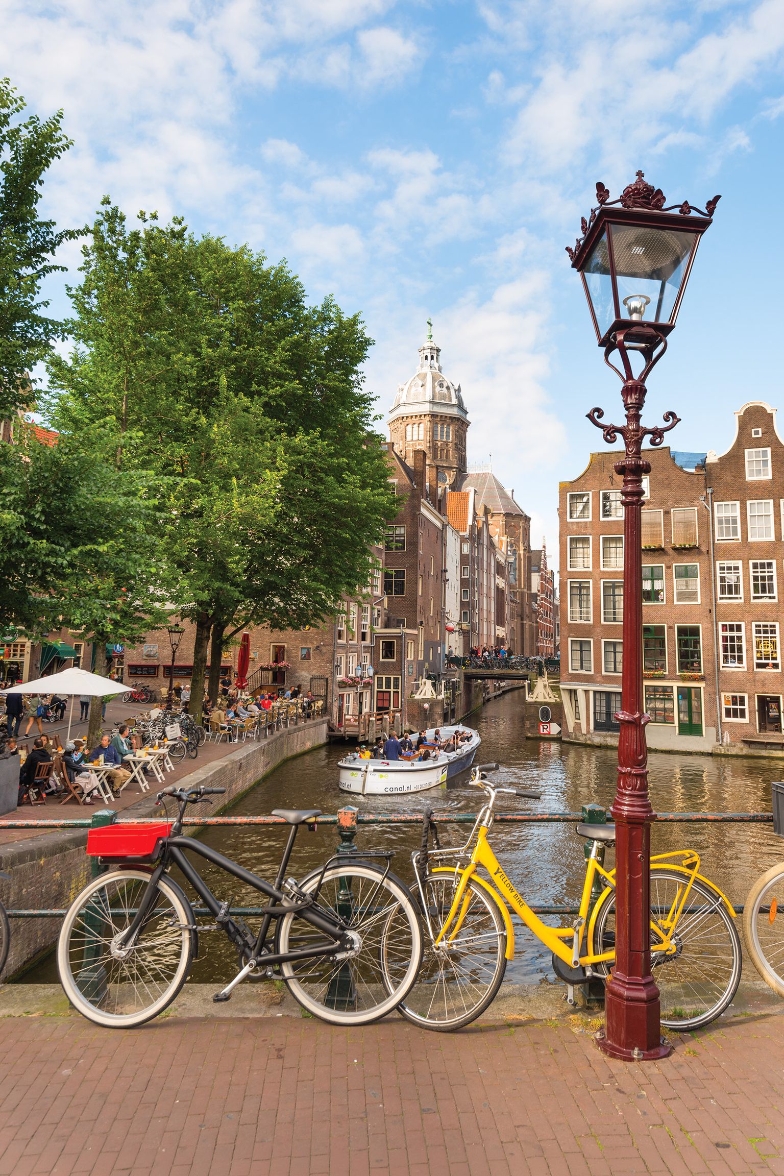 Bicycles parked by a canal in Amsterdam, with a boat passing and traditional buildings in the background under a partly cloudy sky.
