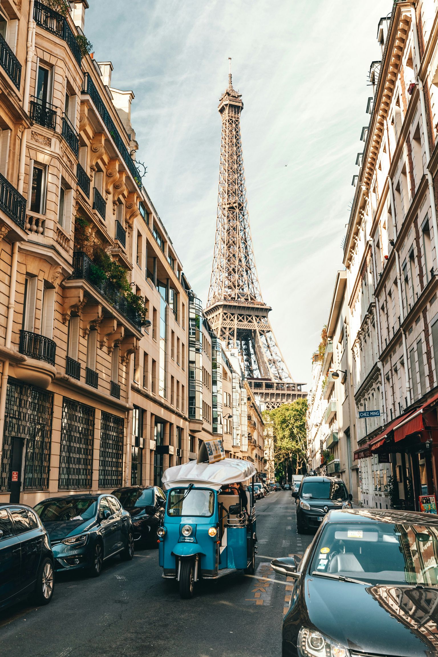 A blue tuk-tuk drives down a narrow Paris street lined with cars and classic buildings, with the Eiffel Tower prominently in the background.