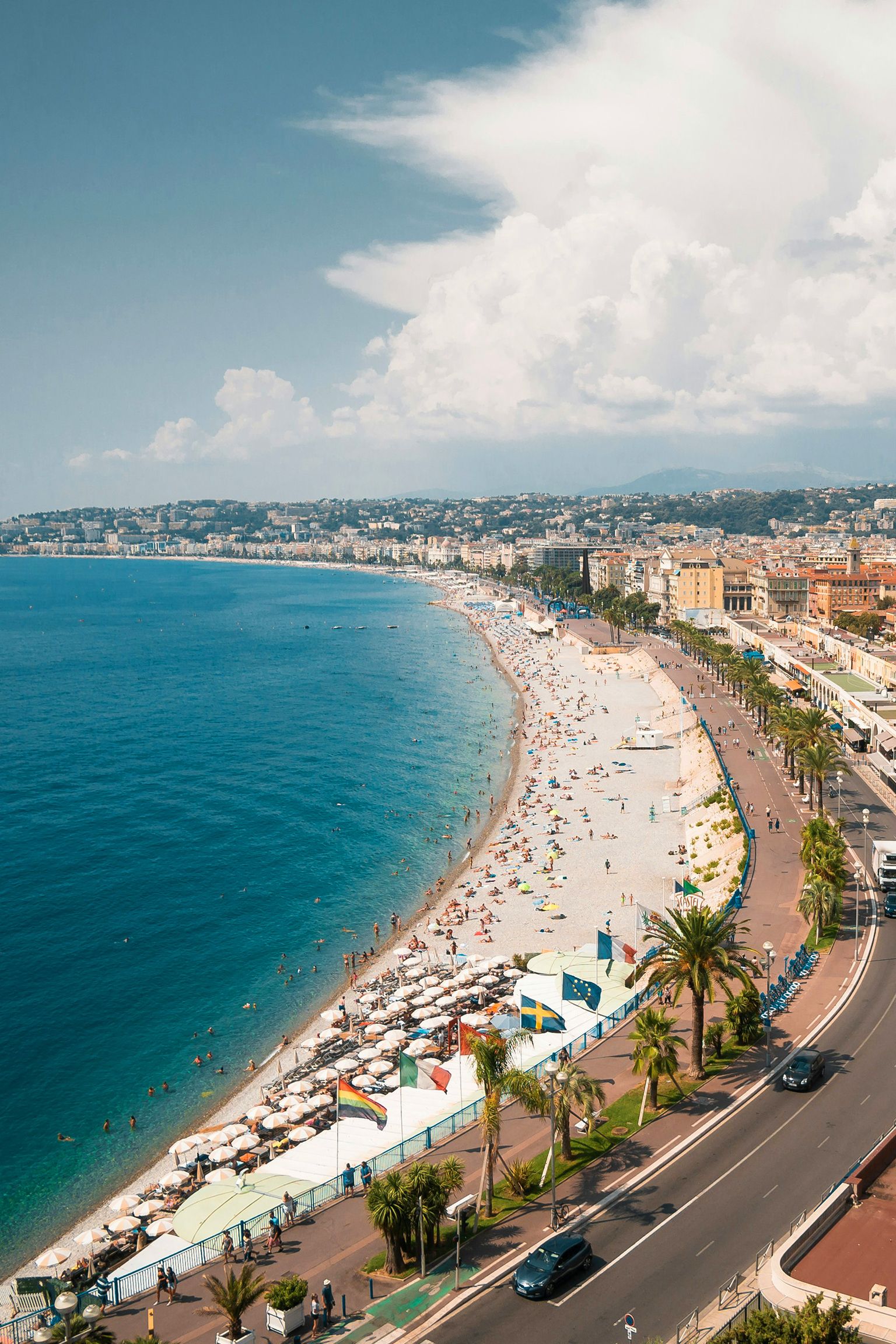 Aerial view of a bustling beach along a curving coastline with turquoise water, vibrant umbrellas, palm trees, and a promenade.