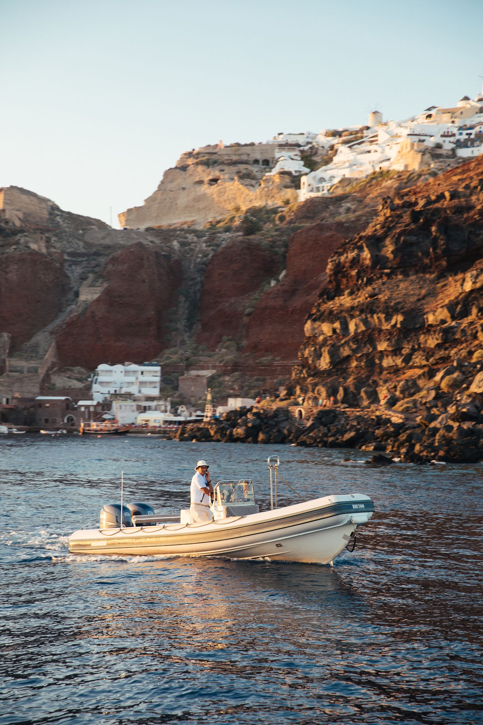 A person navigates a small motorboat on the sea, with rocky cliffs and white buildings on a hillside in the background.