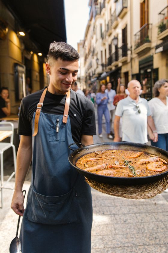 A chef in an apron holds a large pan of paella with shrimp, standing on a busy street with people walking in the background.