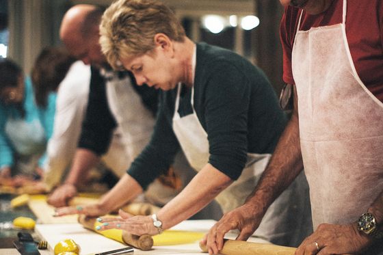People wearing aprons, focused on rolling out yellow dough with rolling pins on a large table in a cooking class setting.