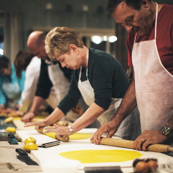 People wearing aprons, focused on rolling out yellow dough with rolling pins on a large table in a cooking class setting.