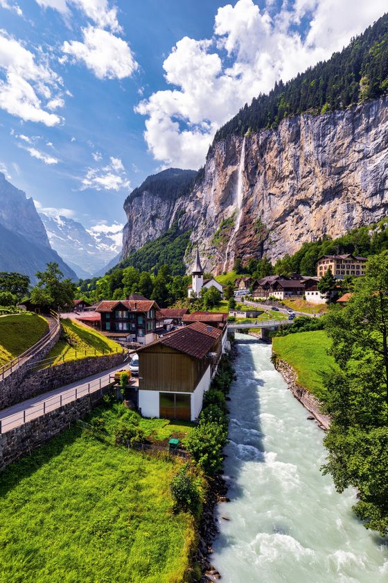 Scenic view of a village with a river, nestled in a lush valley with a waterfall and mountains in the background under a clear blue sky.