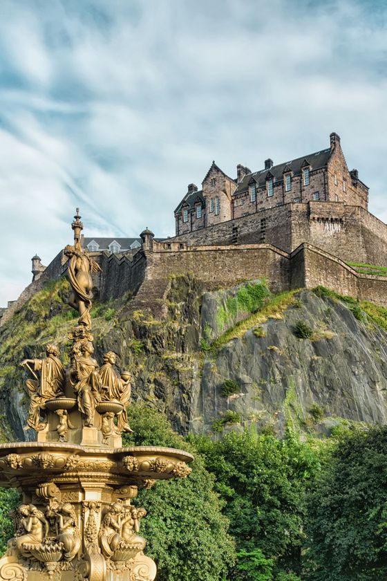 Ornate golden fountain with statues in front of a historic stone castle on a rocky hill, surrounded by green foliage under a cloudy sky.