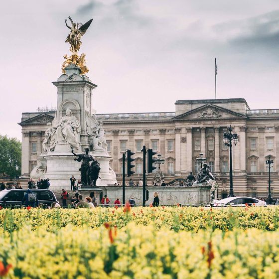 Golden statue atop a white monument with Buckingham Palace in the background, surrounded by a colorful flower garden and people nearby.