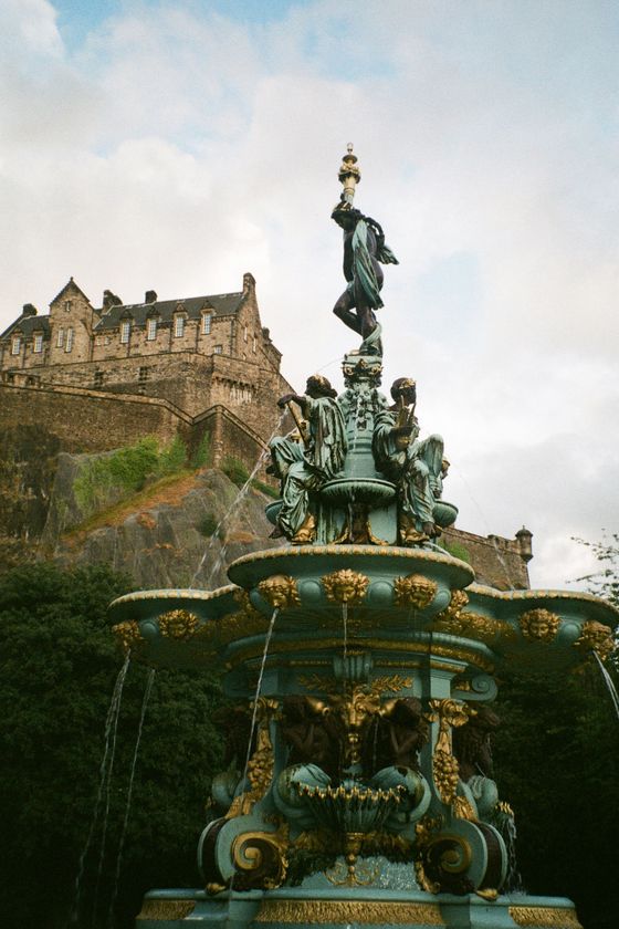 Ornate bronze fountain with figures and water streams, set against a historic stone castle on a grassy hill under a partly cloudy sky.