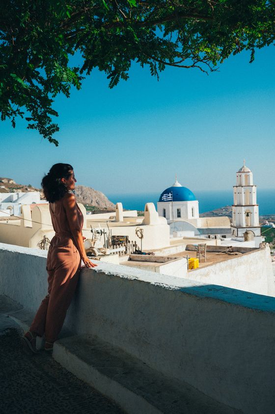 A woman in a peach outfit stands on a terrace, overlooking a Greek island with white buildings and a blue dome under a sunny sky.