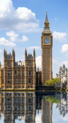 Panoramic view of the Houses of Parliament and Big Ben in London, with reflections in the River Thames under a blue sky with clouds.