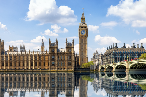 Panoramic view of the Houses of Parliament and Big Ben in London, with reflections in the River Thames under a blue sky with clouds.