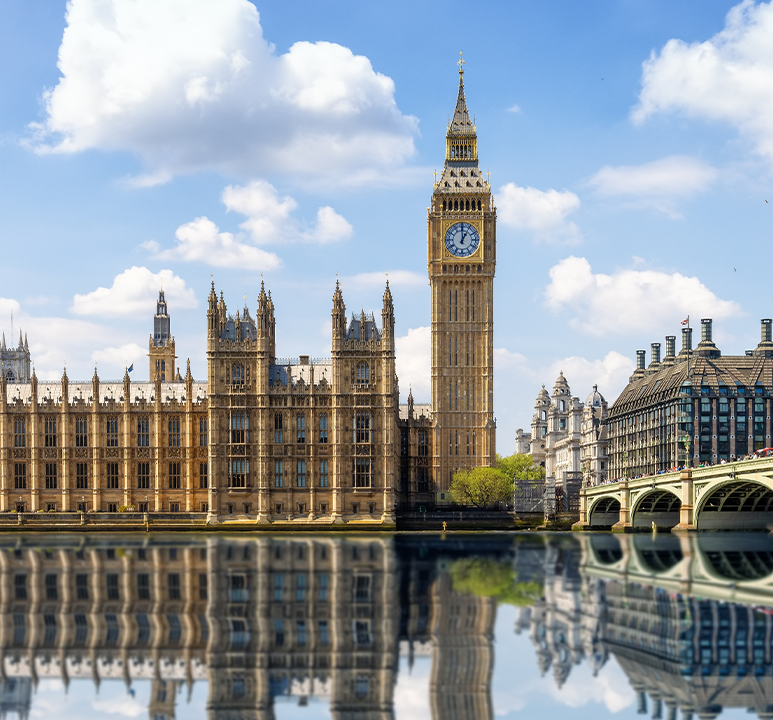 Panoramic view of the Houses of Parliament and Big Ben in London, with reflections in the River Thames under a blue sky with clouds.