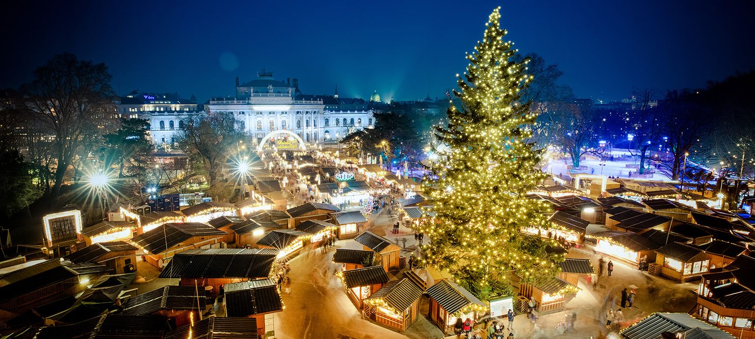 Festive Christmas market at night, with a large illuminated tree and numerous lit stalls, set against a backdrop of a grand building.