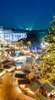 Festive Christmas market at night, with a large illuminated tree and numerous lit stalls, set against a backdrop of a grand building.