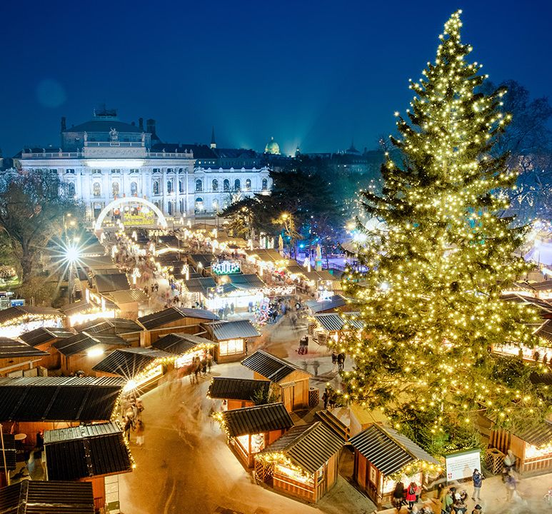 Festive Christmas market at night, with a large illuminated tree and numerous lit stalls, set against a backdrop of a grand building.