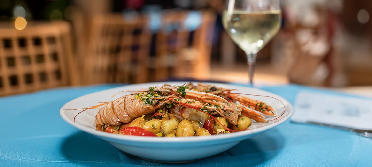 A plate of gnocchi topped with shrimp, herbs, and cherry tomatoes, set on a blue tablecloth with a glass of white wine in the background.
