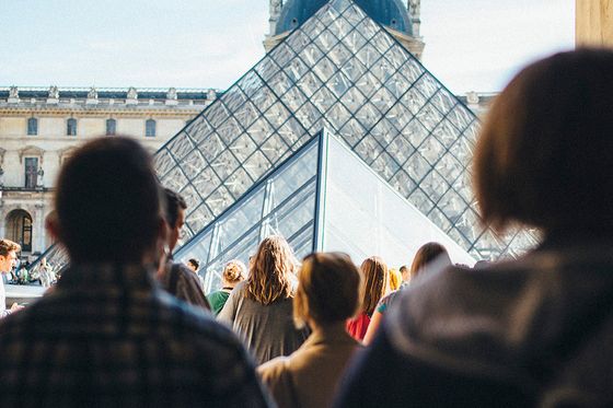 People walking towards the glass pyramid entrance of the Louvre Museum on a sunny day.