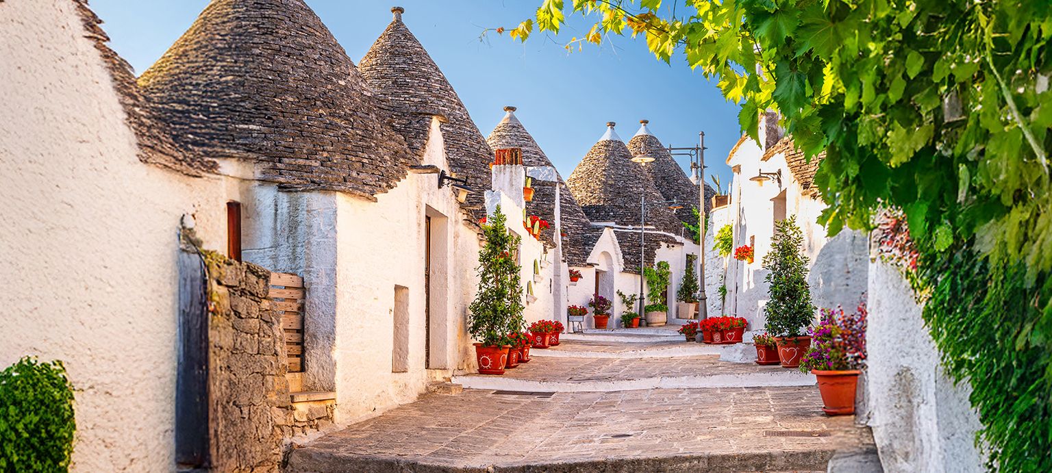 A charming street lined with white trulli houses with conical roofs, adorned with vibrant potted plants, under a clear blue sky.