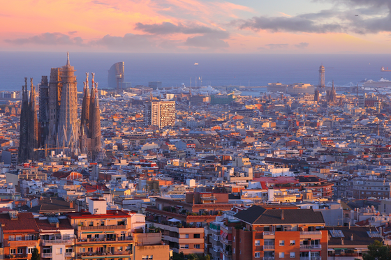Aerial view of Barcelona at sunset, with the iconic Sagrada Familia and city buildings under a pinkish-orange sky.