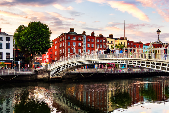 A picturesque bridge arches over a tranquil river, with colorful historic buildings and a vibrant sky in the background.